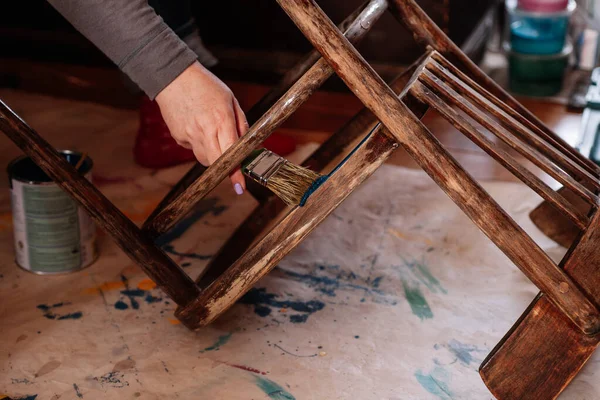 Lady Hand mit Pinsel Malerei Boden des gealterten Stuhls in blauem Kragen in der heimischen Werkstatt für die Neuerfindung von Möbeln. Wiederverwendung alter Dinge. Zukunft durch nachhaltiges Handeln denken Stockfoto