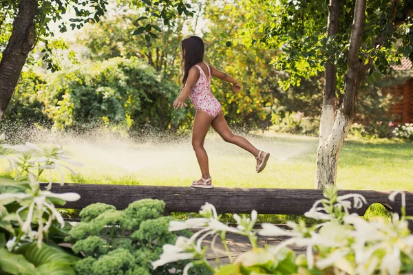 Das weibliche Kind hat Spaß dabei, auf einem Fuß das Gleichgewicht zu finden und sich tagsüber mit Wassersprengern, Haus und grünen Bäumen im Hintergrund zu vergnügen. Träumen davon, Turner zu werden lizenzfreie Stockbilder