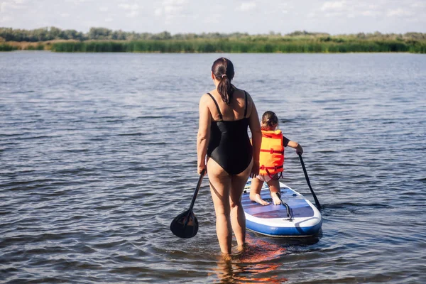 Mãe caucasiana de meia idade cuidando da filha pequena sup embarque no lago com remo nas mãos em colete salva-vidas. Férias activas cheias de aventuras. Inculcação de amor por esportes desde a infância — Fotografia de Stock