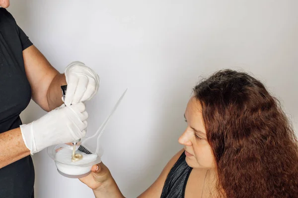 Las manos femeninas en guantes de látex exprimiendo el contenido del tubo de teñido del cabello en un tazón en la mano de una mujer pelirroja mirando la sustancia colorante sobre un fondo blanco —  Fotos de Stock