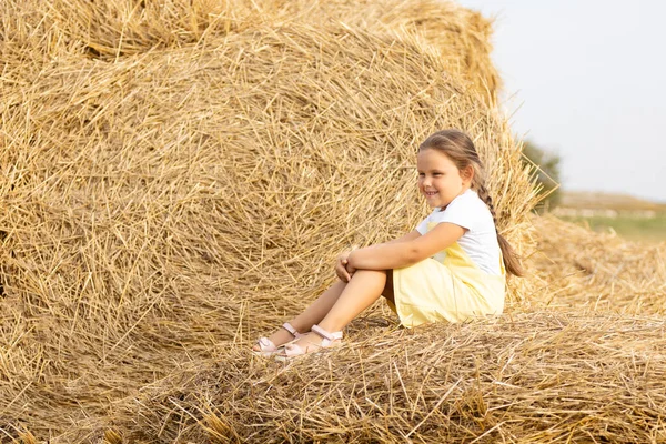 Portrait of happy little girl with eyes squinted from sun and long braid on head looking away sitting on haystacks. Time away from city in country field with tons of hayricks Royalty Free Stock Photos