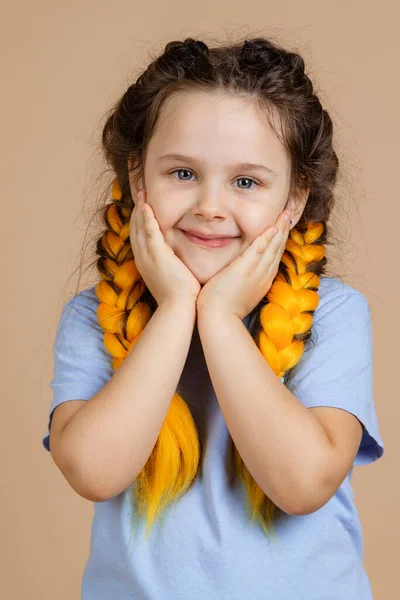 Retrato de niño pequeño con las manos en las mejillas ligeramente sonriente mirando a la cámara con ojos brillantes con trenzas de kanekalon amarillo en la cabeza sobre fondo beige con camiseta azul y jeans — Foto de Stock