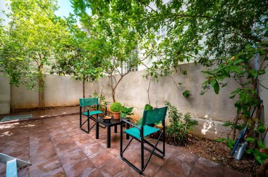 Arka bahçe terası fayanslı zemin ve bahçe mobilyaları, iki yeşil sandalye ve dekorlu ince siyah masa, ağaçlar ve beyaz duvar. Yaz güneşli bir gün.