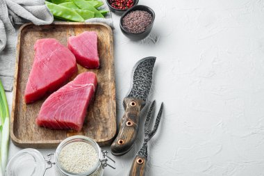 Çiğ ton balığı bifteği, taze kırmızı ton balığı fileto, yeşil bezelye, susam ve baharat seti, ahşap tepside, beyaz taş zemin üzerinde, kopyalanacak yer ve metin için yer var.