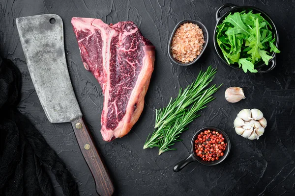 Cooking meat background. Raw aged beef t-bone steak, with spices and herbs for grill set, on black stone background, top view flat lay