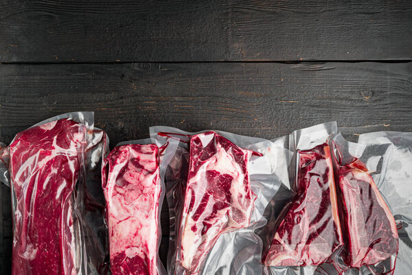 Vacuum packed organic raw beef classic cuts set, tomahawk, t bone, club steak, rib eye and tenderloin cuts, on black wooden table background, top view flat lay, with copy space for text