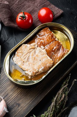 Tütsülenmiş somon filetosu konserve balık seti, ahşap kesme tahtası, siyah arka planda bitkiler ve malzemeler.