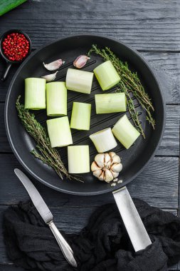 Çiğ pırasa, ızgara tavada doğranmış oninon, otlarla pişirilmemiş, siyah ahşap masa manzaralı.