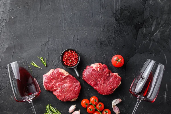 Raw rump steaks from organic beef meat cuts near red wine glasses with rosemary, garlic and spices over black textured  background, top view with space for text