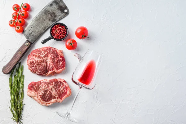 Organic top blade steak, raw beef meat with seasonings, rosemary, garnet, rose wine glass and butcher cleaver. White textured background. Top view with space for text