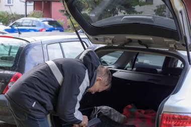 Orta yaşlı bir adam bagajı açık bir arabanın yanında duruyor ve içine bakıyor, şehir manzarası.