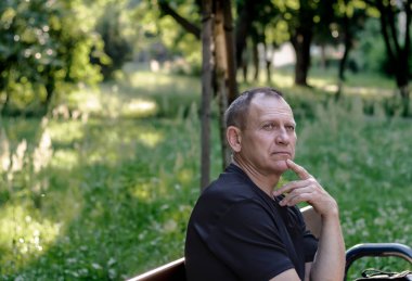 Olgun, cesur bir adamın portresi. 60 yaşında, siyah tişörtlü, bir bankta oturuyor, doğanın arka planına karşı.