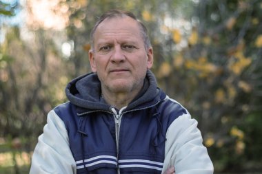Erkek, olgun, orta yaşlı, 60 yaşında, kır saçlı, kırışıklıkları olan, arka planda bel hizasında portresi olan bir adam.