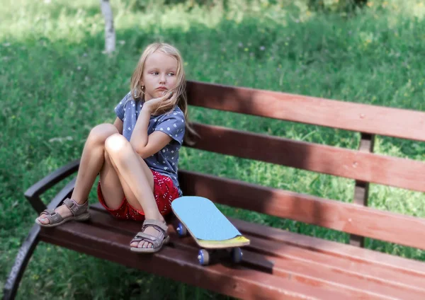 小さな悲しい女の子 ブロンド6 7歳は スケートボードの横にある高価な外観のベンチに公園に座っています — ストック写真