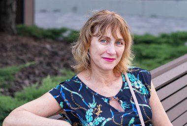 Güzel, sarışın, orta yaşlı bir kadının portresi. 50-55 yaşlarında, elbise giymiş, parkta bankta oturuyor.