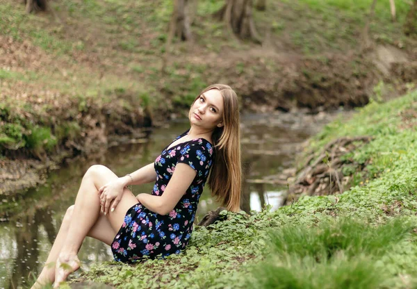 長い髪をした若い美しい細いヨーロッパの女の子が川のほとりの森の中に座っています — ストック写真