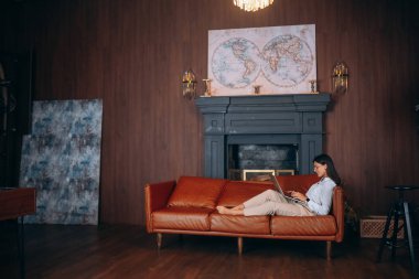 Genç bir kadın dizüstü bilgisayarda çalışırken kırmızı bir koltukta oturuyor..