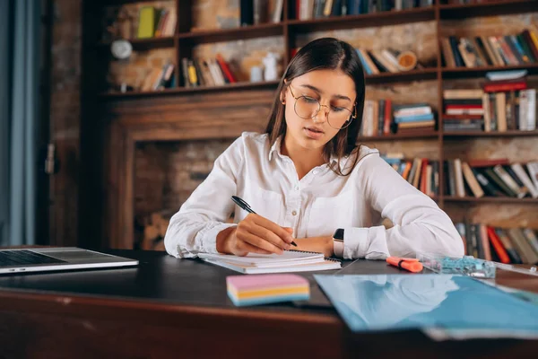Mladá žena v brýlích píše do zápisníku, zatímco sedí u stolu — Stock fotografie