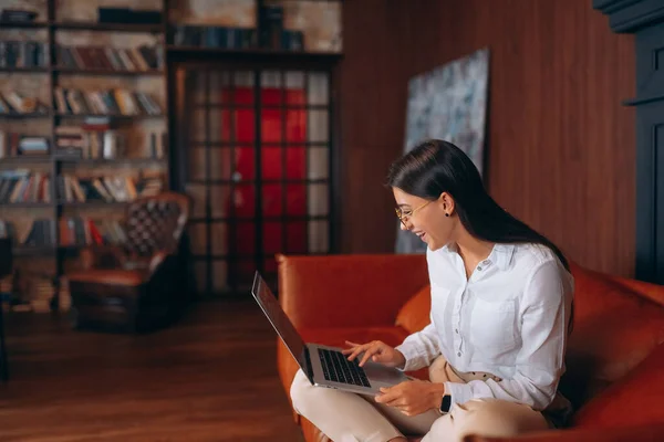 Giovane donna si siede su un divano rosso mentre lavora in un computer portatile. — Foto Stock