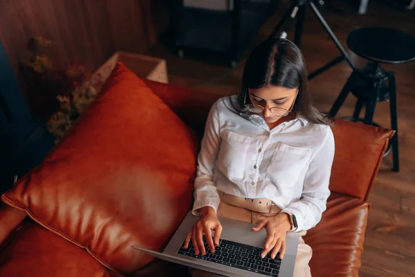Giovane donna si siede su un divano rosso mentre lavora in un computer portatile. — Foto Stock