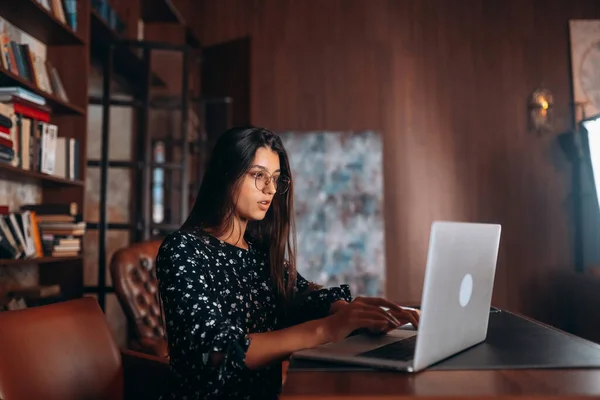 Молодая женщина в очках работает за ноутбуком, сидя за столом — стоковое фото