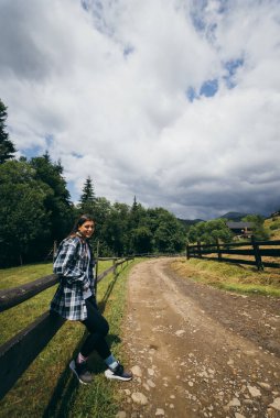 Genç, çekici, beyaz bir kadın çitin yanında duruyor.