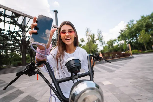 Menina sentada em uma scooter e mostrando o telefone para a câmera — Fotografia de Stock
