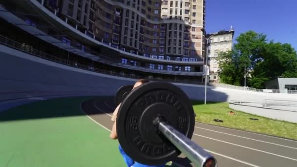 Молодий хлопець піднімає бар на стадіоні, тренування на відкритому повітрі — стокове відео