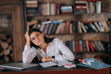 Ofisteki genç kadın, kitaplıkların arka planındaki sorun üzerine düşünüyor.