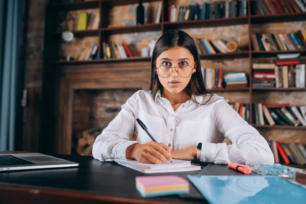 Junge Frau mit Brille schreibt in Notizbuch, während sie am Tisch sitzt — Stockfoto