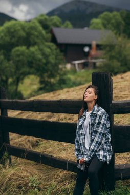 Genç, çekici, beyaz bir kadın çitin yanında duruyor.