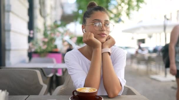 Hermosa joven sentada en la cafetería de la calle y escuchando música — Vídeo de stock