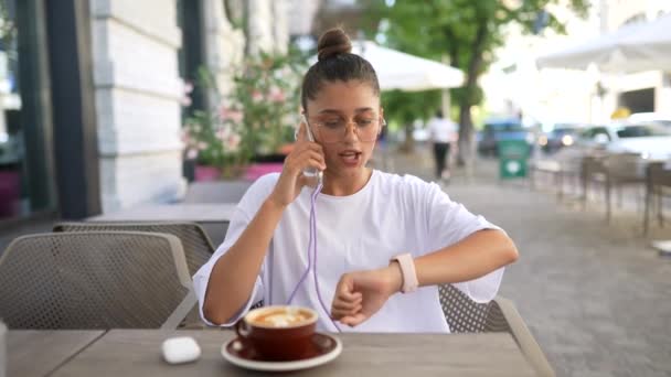 Hermosa joven sentada en la cafetería de la calle hablando en un teléfono inteligente — Vídeo de stock