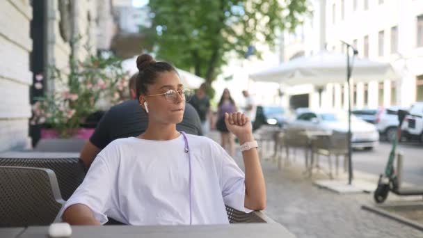 Gyönyörű fiatal nő ül street cafe és zenét hallgatni — Stock videók