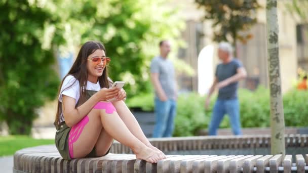 Mujer bastante joven utilizando la aplicación de teléfono móvil en el parque de la ciudad — Vídeos de Stock