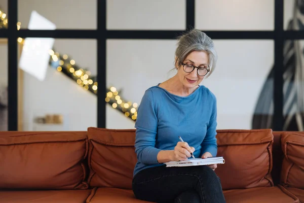 Ευτυχισμένη ηλικιωμένη γυναίκα με γυαλιά που γράφει στο σημειωματάριο στο σπίτι — Φωτογραφία Αρχείου