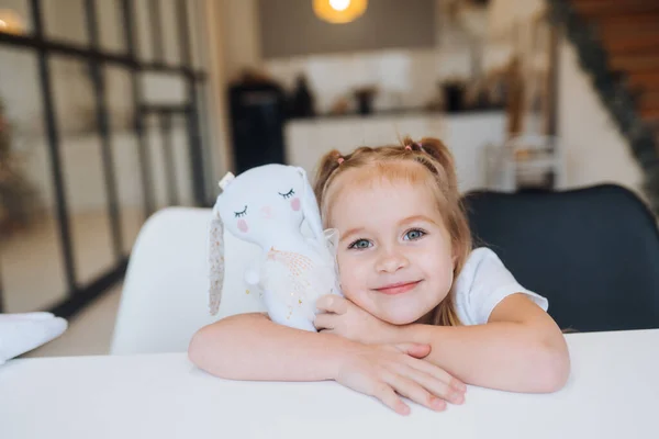 Little uśmiechnięta dziewczyna przytula ulubione zabawki siedząc przy stole — Zdjęcie stockowe