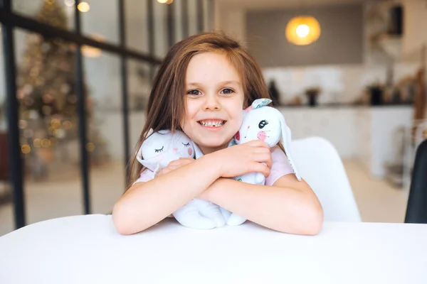 Kleines lächelndes Mädchen umarmt Lieblingsspielzeug, während es am Tisch sitzt — Stockfoto