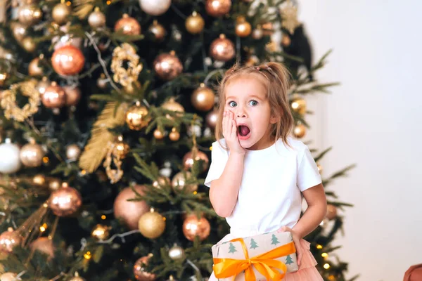 Чарівна маленька здивована дівчинка тримає подарунок на тлі ялинки — стокове фото