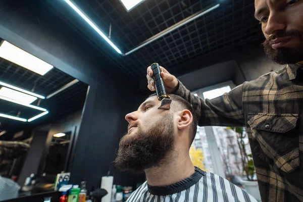 O barbeiro trabalha com um cortador de barba. Hipster cliente recebendo corte de cabelo. — Fotografia de Stock