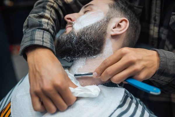 Young bearded man getting shaved by hairdresser at barbershop — Stock Photo, Image