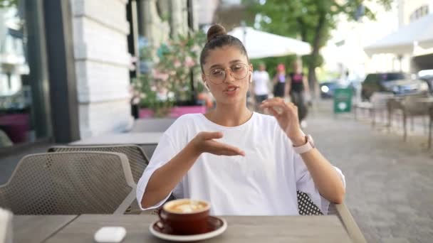 Chica está sentado en la cafetería al aire libre y habla con la cámara — Vídeo de stock