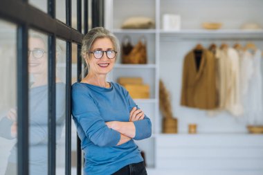 Yaşlı bir kadın, kollarını göğsünde kavuşturup kendinden emin bir gülümsemeyle kameraya bakıyor.