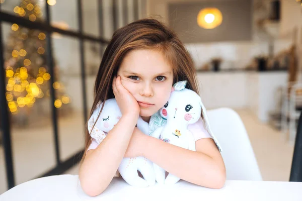 Маленькая грустная девочка обнимает любимую игрушку, сидя за столом — стоковое фото