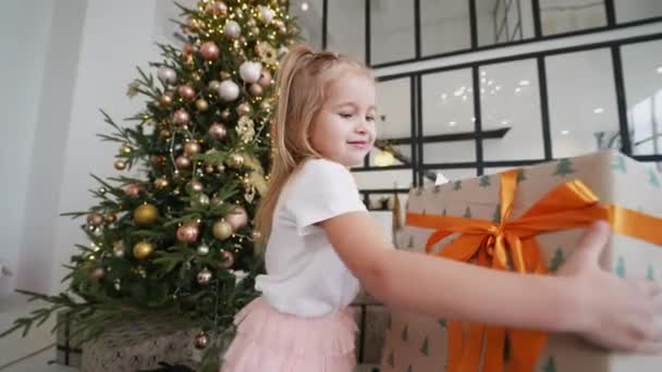 チャーミングな女の子はクリスマスツリーの背景に贈り物を保持しています — ストック動画