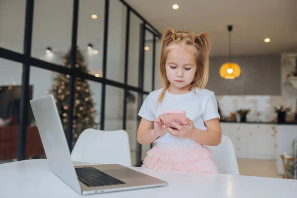 Küçük çocuk dizüstü bilgisayarlı cep telefonuyla masada oturuyor. — Stok fotoğraf