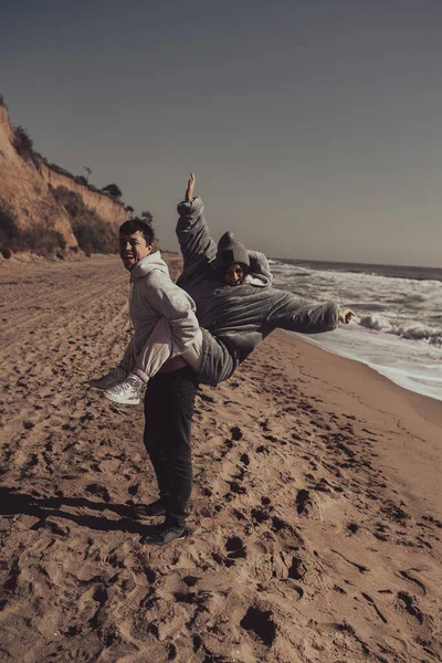 Hombre lleva a su novia en la espalda, pareja divirtiéndose en la orilla del mar — Foto de Stock