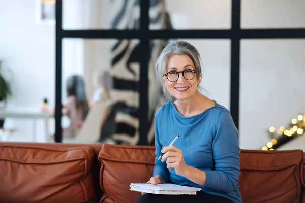 Glückliche Seniorin mit Brille schreibt zu Hause ans Notizbuch — Stockfoto