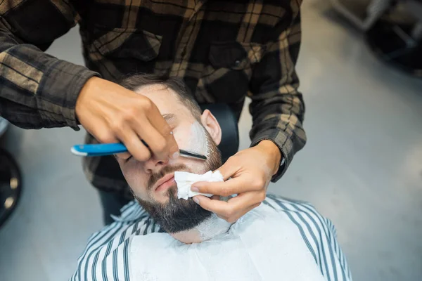 Молодой бородатый мужчина сбривается парикмахером в парикмахерской — стоковое фото