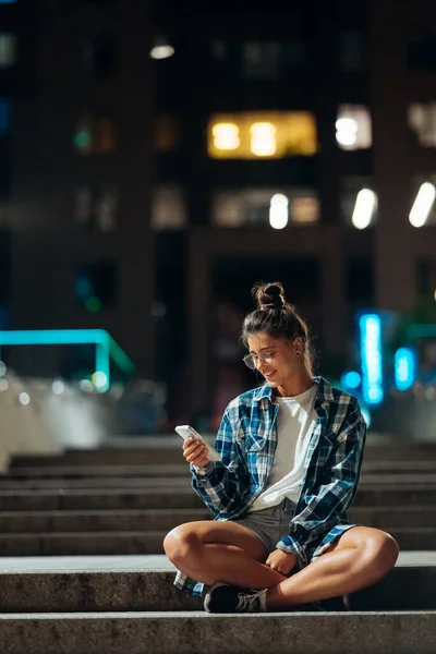 Akıllı telefonlu bir kadın, gece şehrinde uzun taş basamaklarda oturur. — Stok fotoğraf